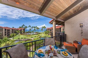 A balcony with a table set for dining, overlooking a resort with palm trees and a view of the ocean.