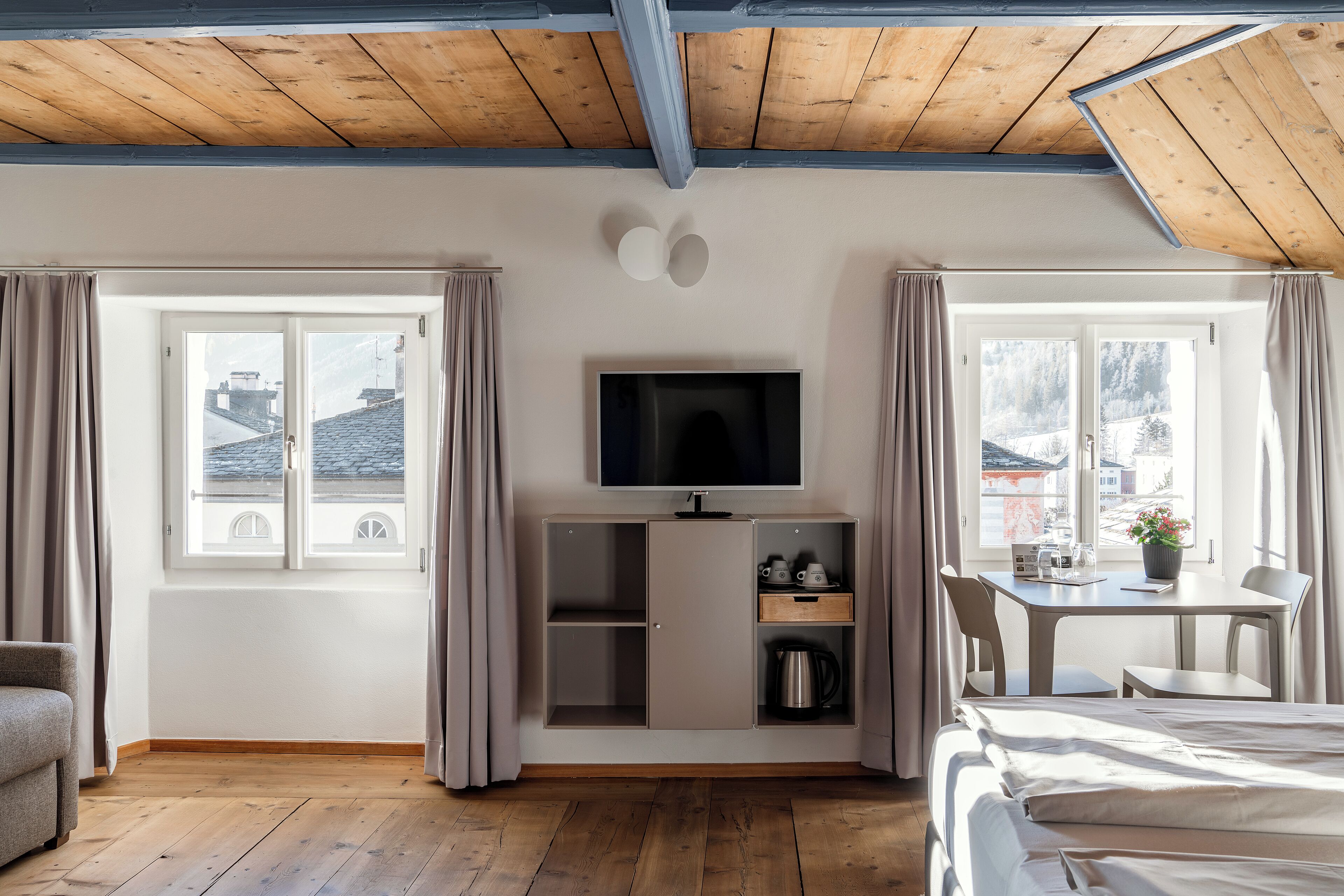 A hotel room with a wooden ceiling, a TV on a cabinet, a bed, a desk with a chair, and a window with curtains.