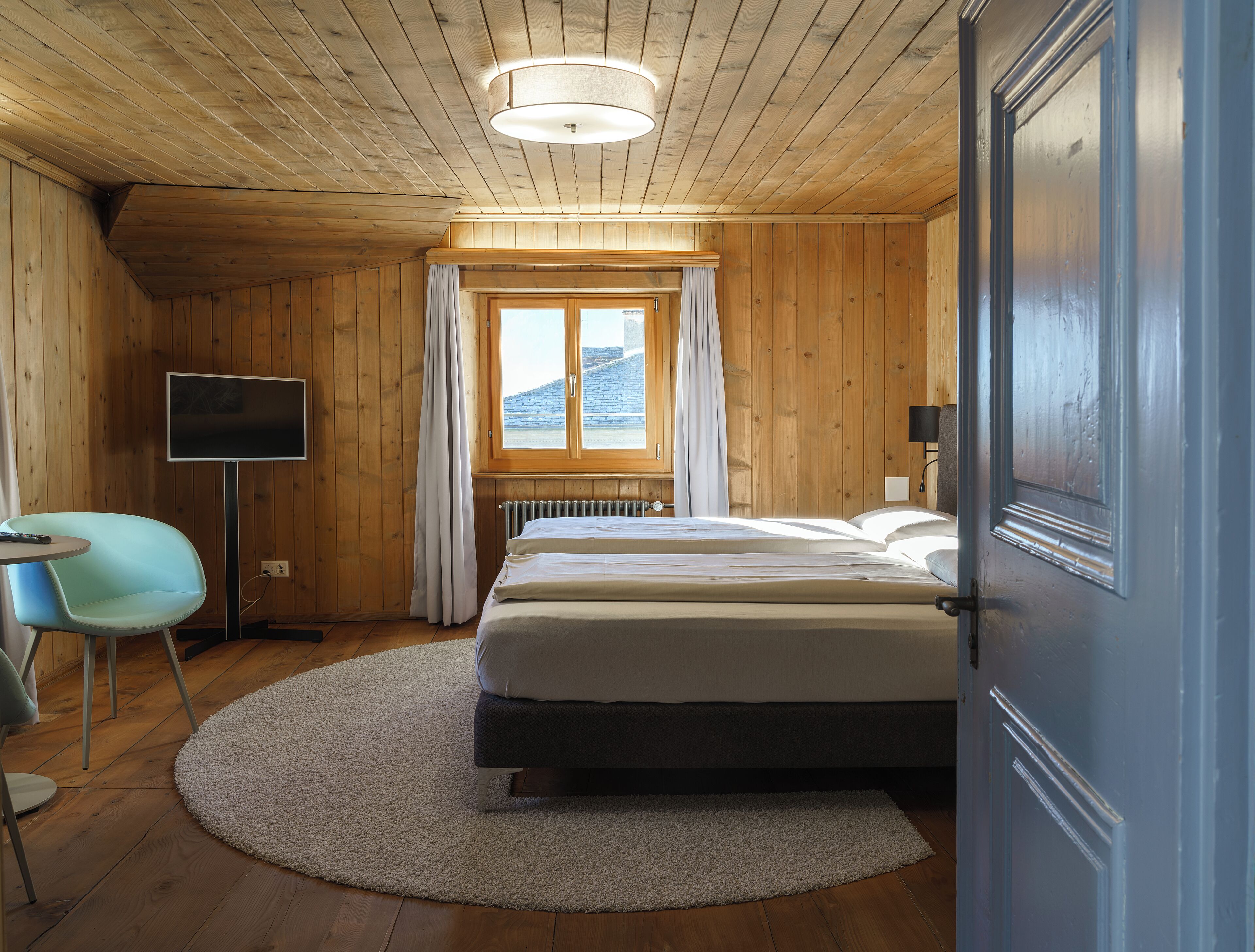 A wooden-paneled room with a bed, a television, a chair, and a window with curtains.