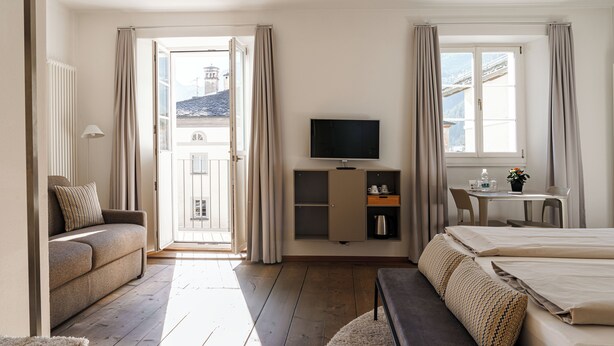 A hotel room with a sofa, a bed, a TV, a desk, and a view of the outside. - Cà del Borgo, Rooms & Suites (Poschiavo)