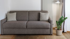 A grey sofa with patterned cushions, a potted plant, and a floor lamp in a room with wooden flooring and vertical blinds. - Cà del Borgo, Rooms & Suites (Poschiavo)