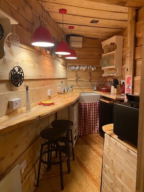 A cozy kitchen with wooden walls, a wooden countertop, two bar stools, a sink, and hanging red pendant lights. - Unique and Bespoke Stunning Thatched Dorset Shepherds Huts with Private Hot Tub (Dorchester)