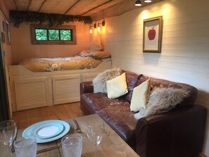 A cozy room with a bed, a leather sofa, a dining table with plates and glasses, and a framed picture on the wall. - Unique and Bespoke Stunning Thatched Dorset Shepherds Huts with Private Hot Tub (Dorchester)
