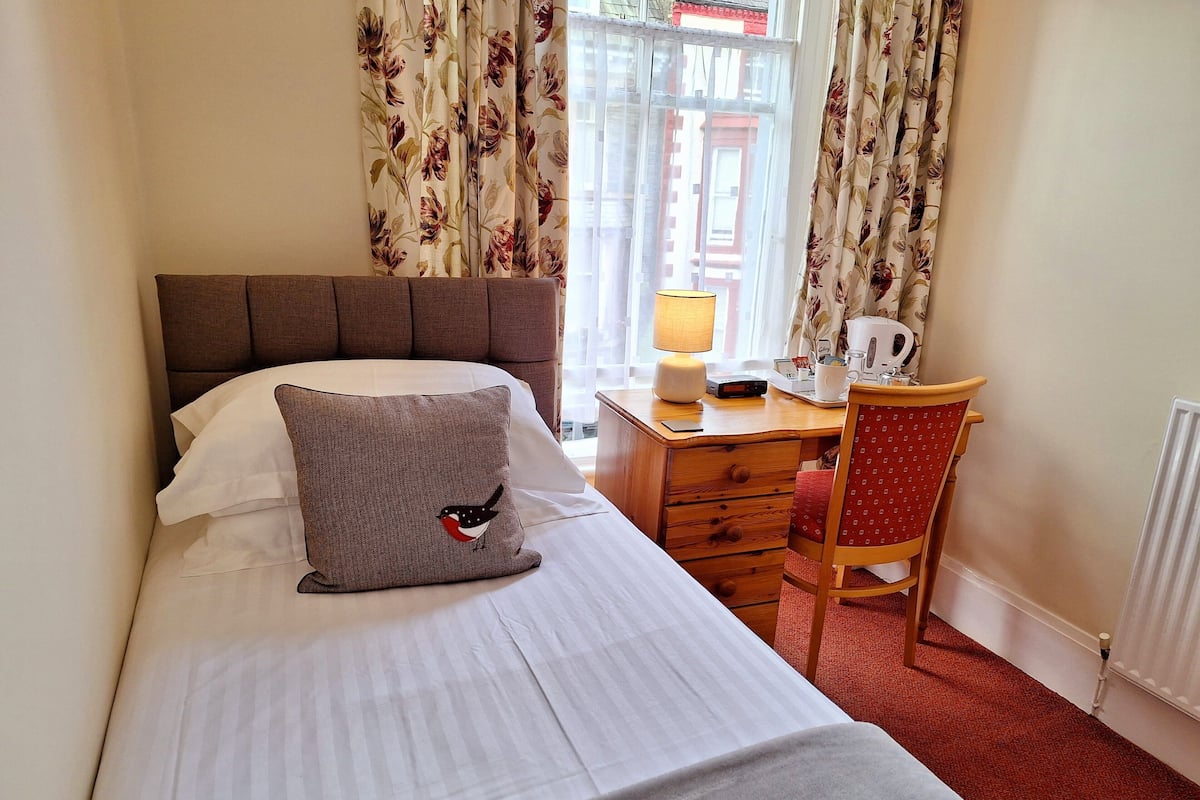 A neatly made bed with a pillow, a wooden desk with a lamp, a chair, and a radiator.