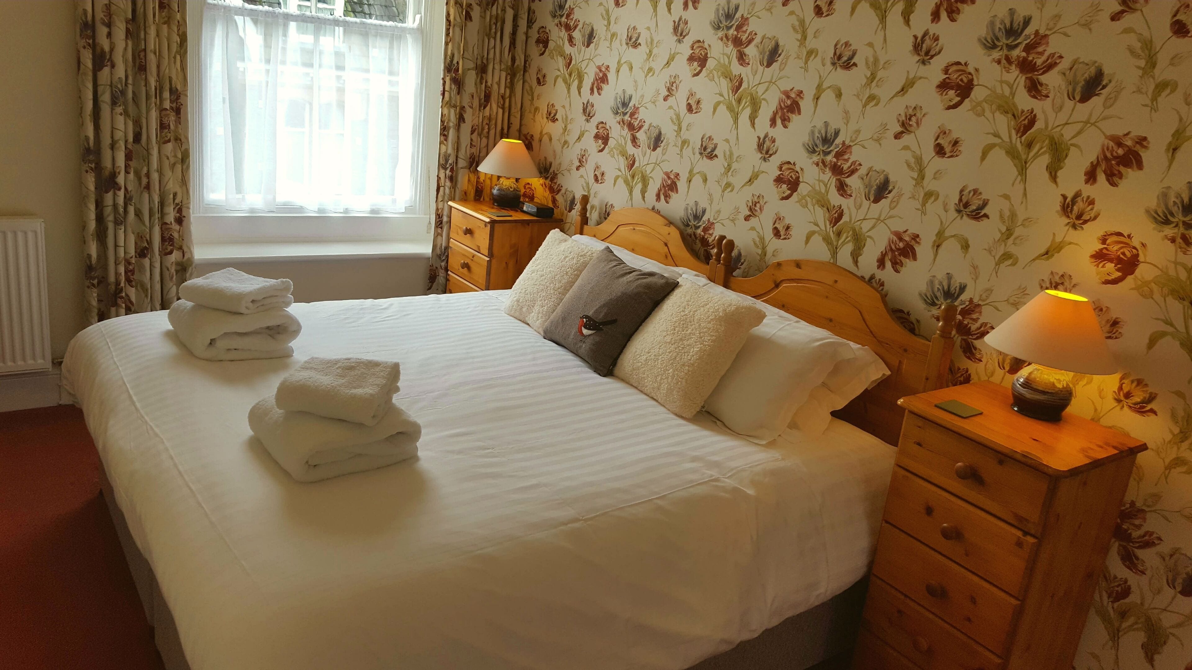 A neatly made bed with white linens and a stack of folded towels. There is a wooden nightstand with a lamp and a small drawer. The room has floral wallpaper and a window with curtains.