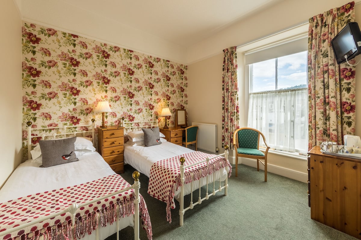 A bedroom with two beds, a desk, a chair, a TV, and a window with curtains.