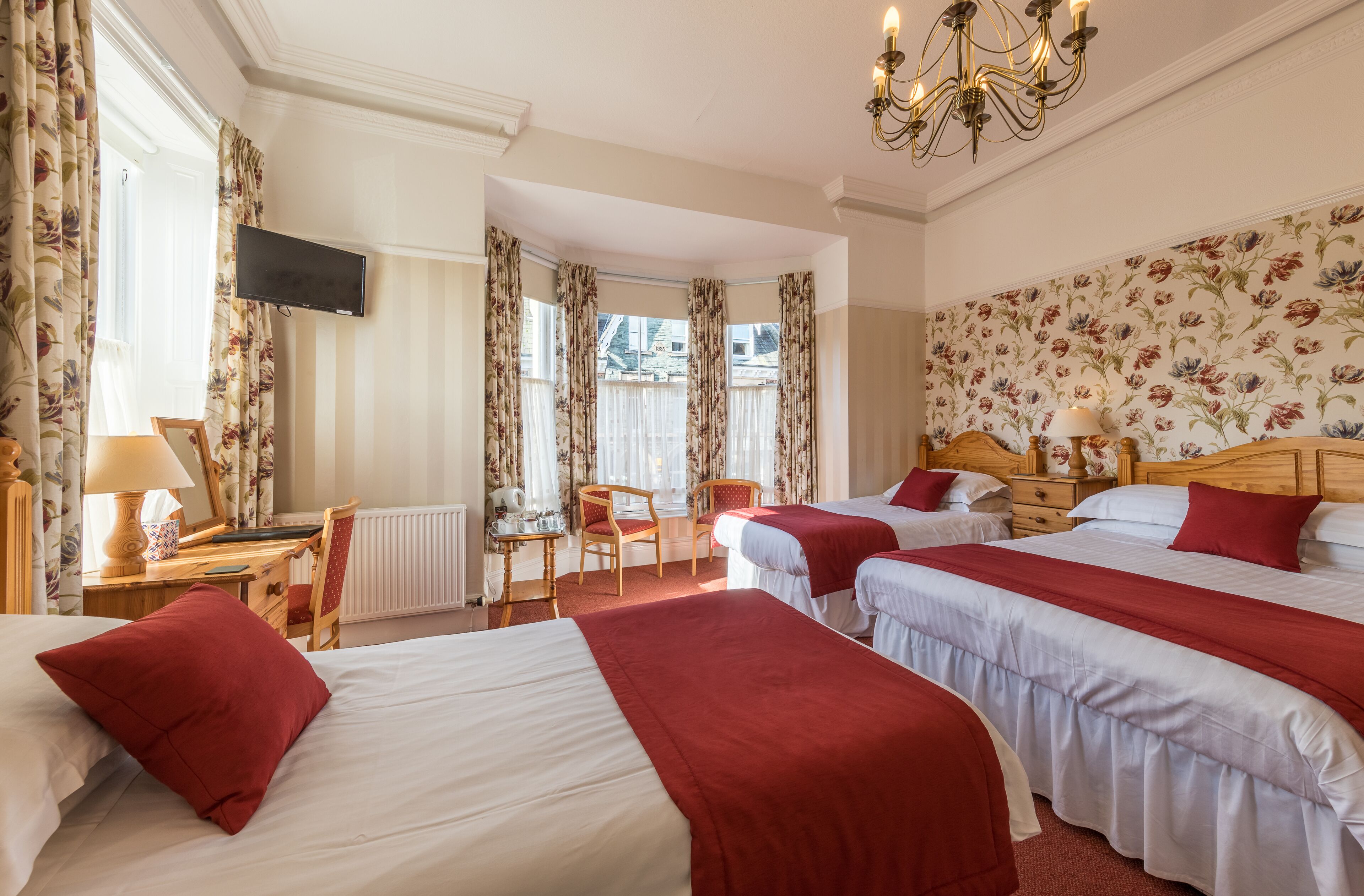 A hotel room with three beds, a desk with a chair, a TV, and a chandelier.