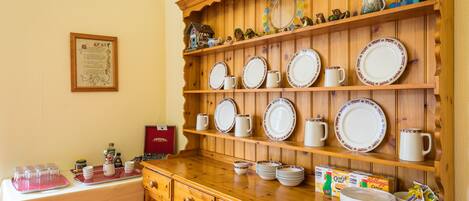 A kitchen area with wooden shelves holding plates, mugs, and a decorative mirror. There are also various kitchen items and a red box on the counter.