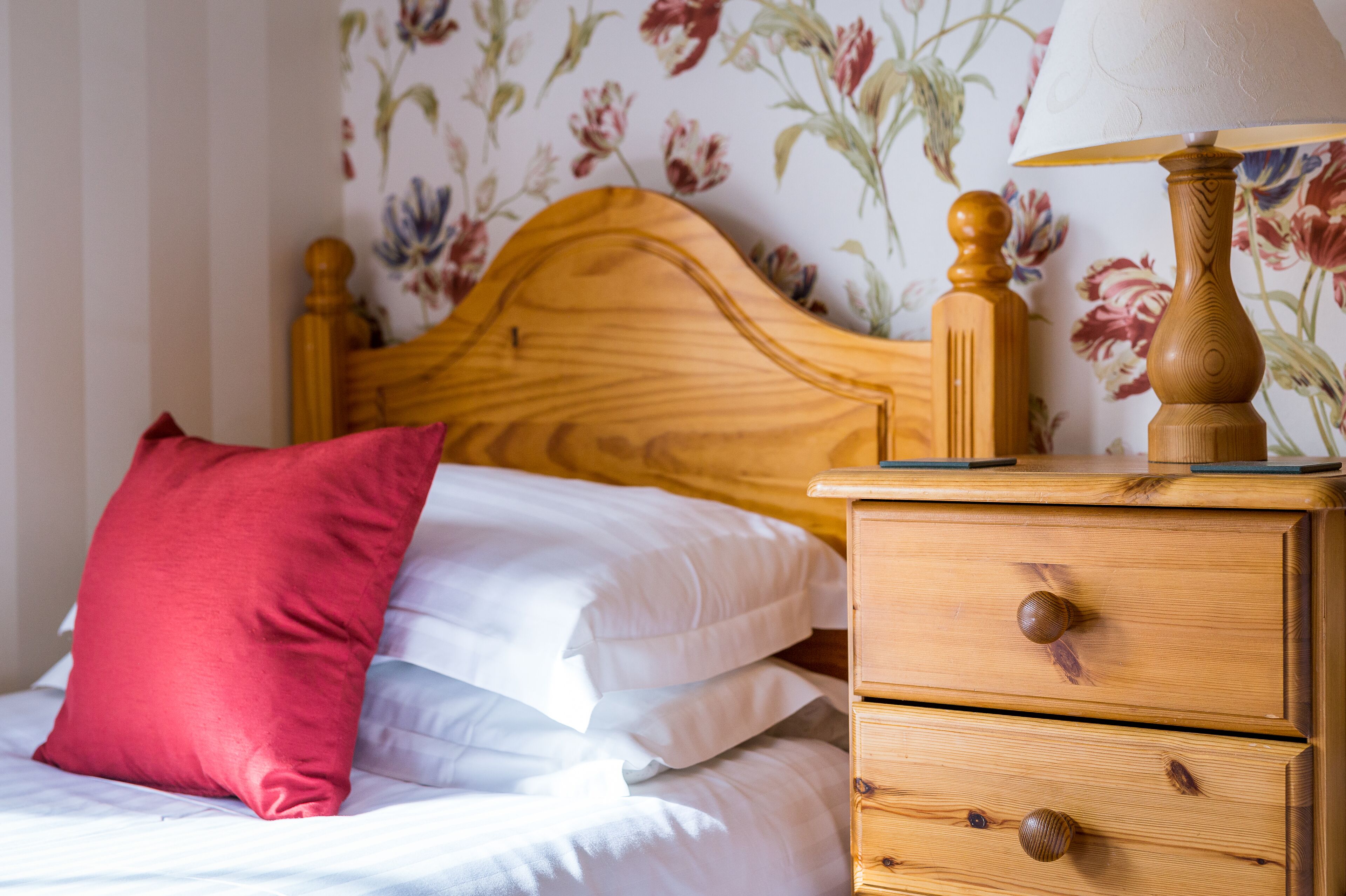 A wooden bed with white and red pillows, a wooden nightstand with drawers, and a lamp.