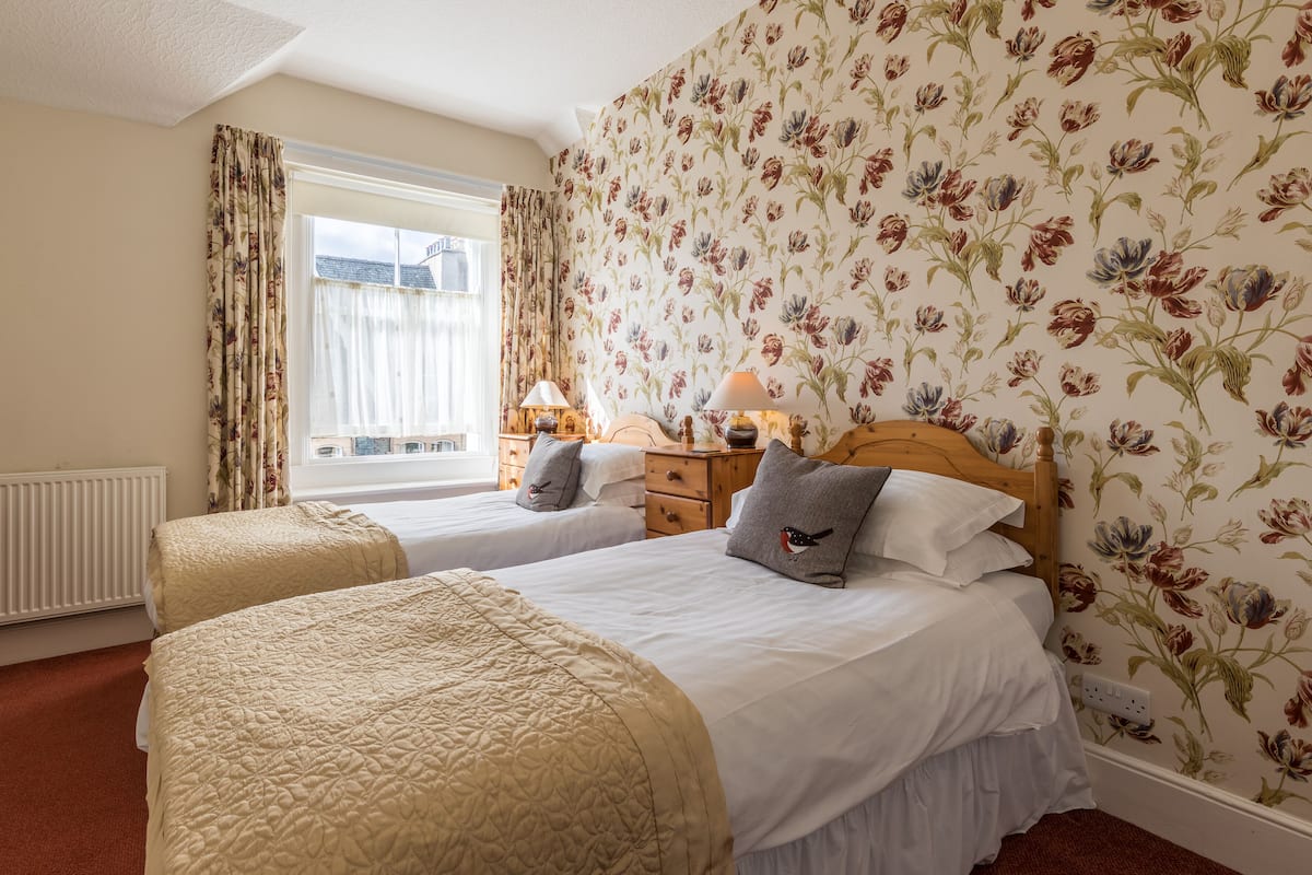 A room with two beds, floral wallpaper, a window with curtains, a bedside table with a lamp, and a radiator.