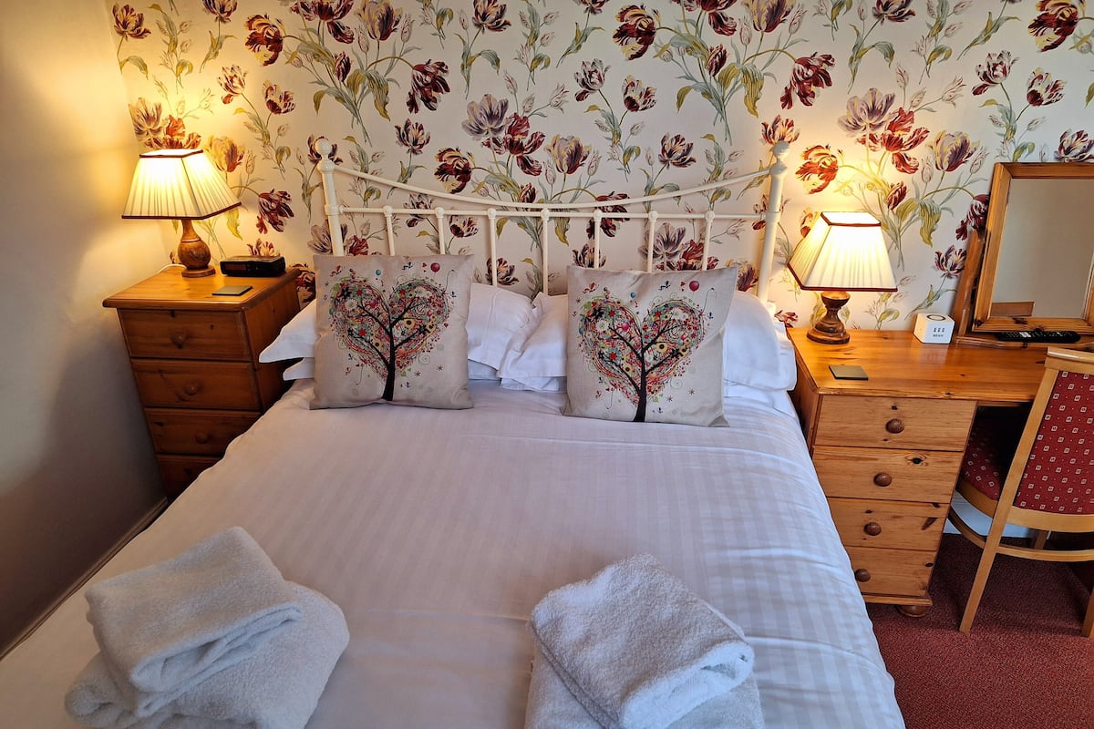 A neatly made bed with two towels, a wooden dresser, a desk with a lamp, and floral wallpaper.
