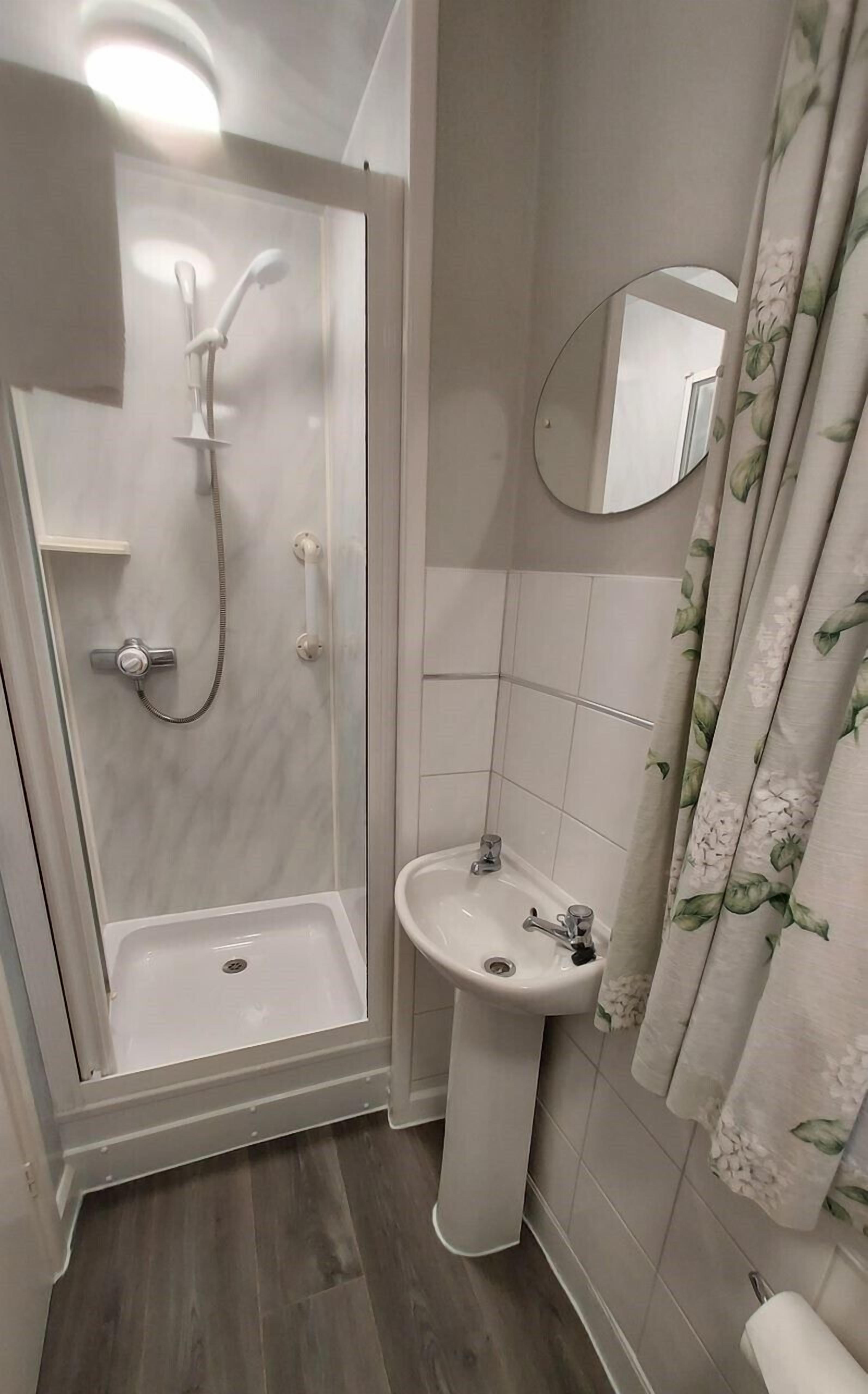 Une salle de bain avec une douche à l’italienne, un lavabo sur colonne, un miroir et des rideaux à motifs floraux.