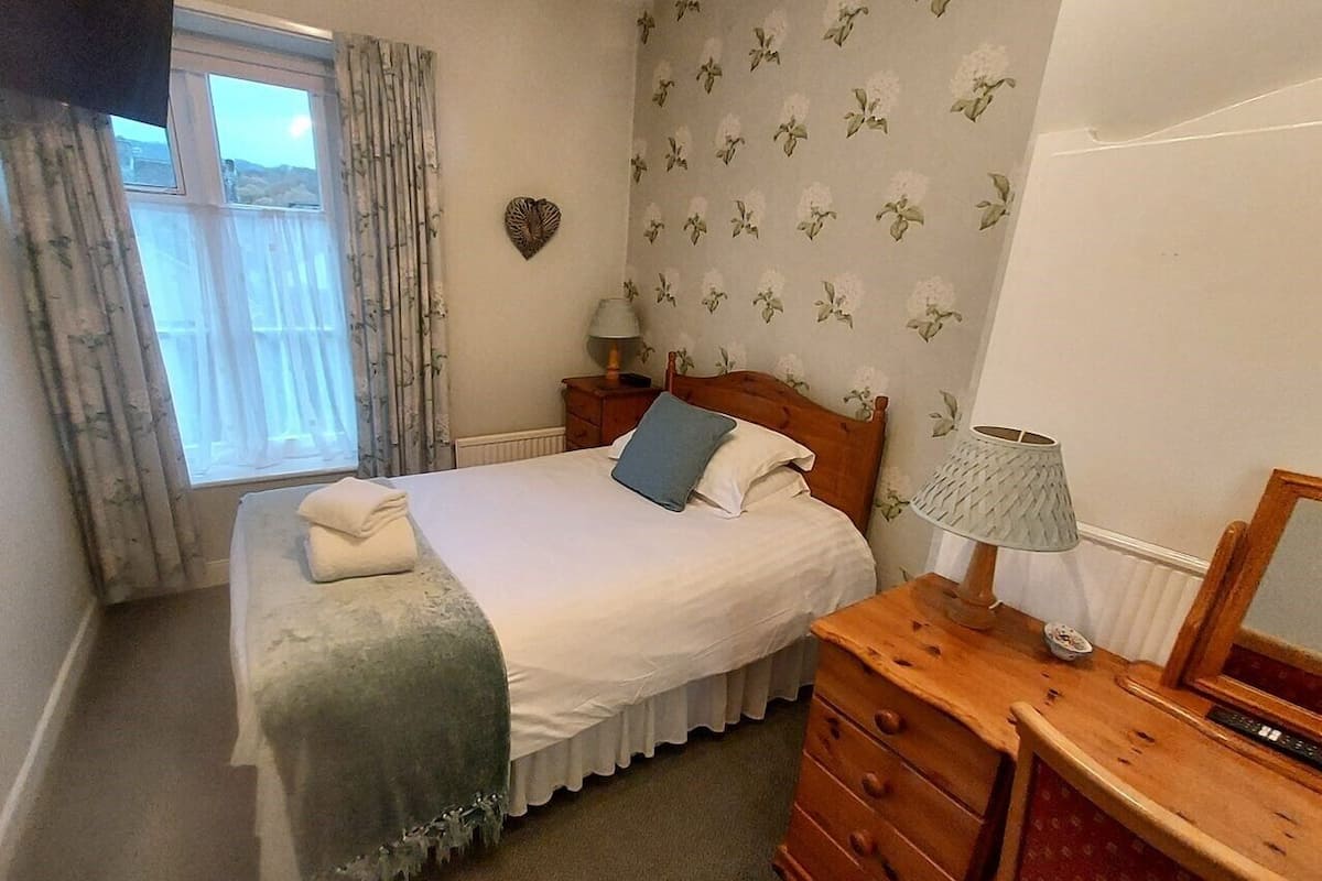 A bedroom with a bed, a dresser, a nightstand, a lamp, and a window with curtains.