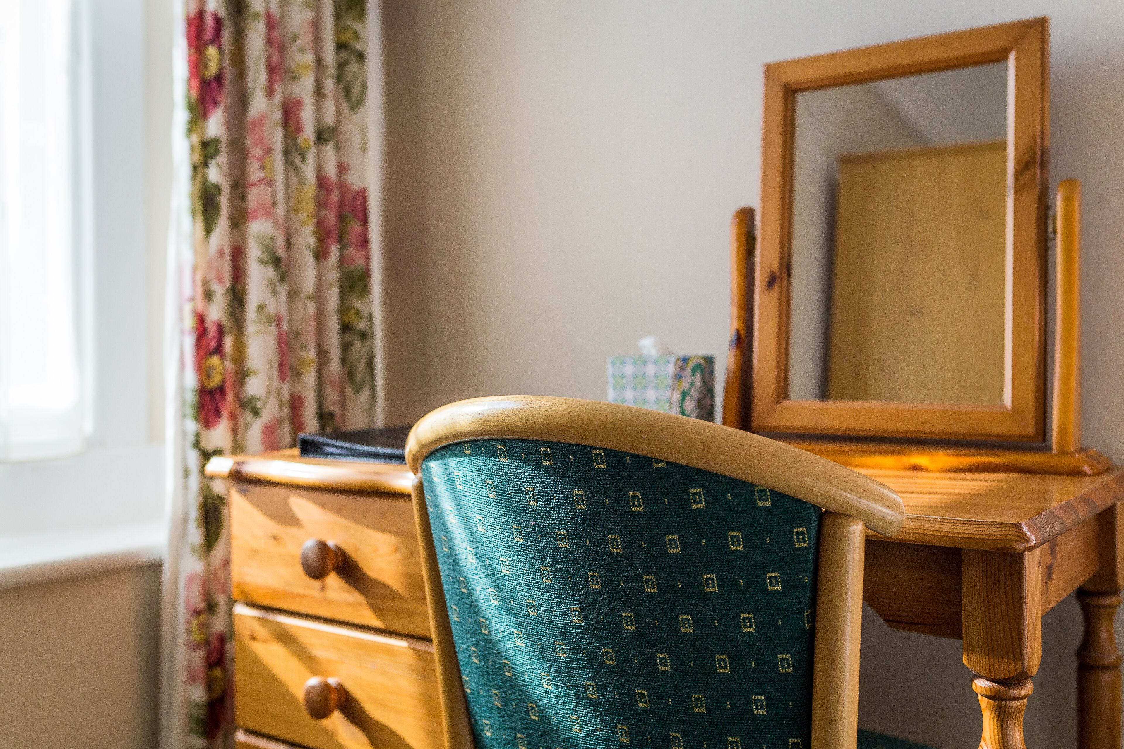A wooden desk with a mirror, a teal chair, and a floral curtain.