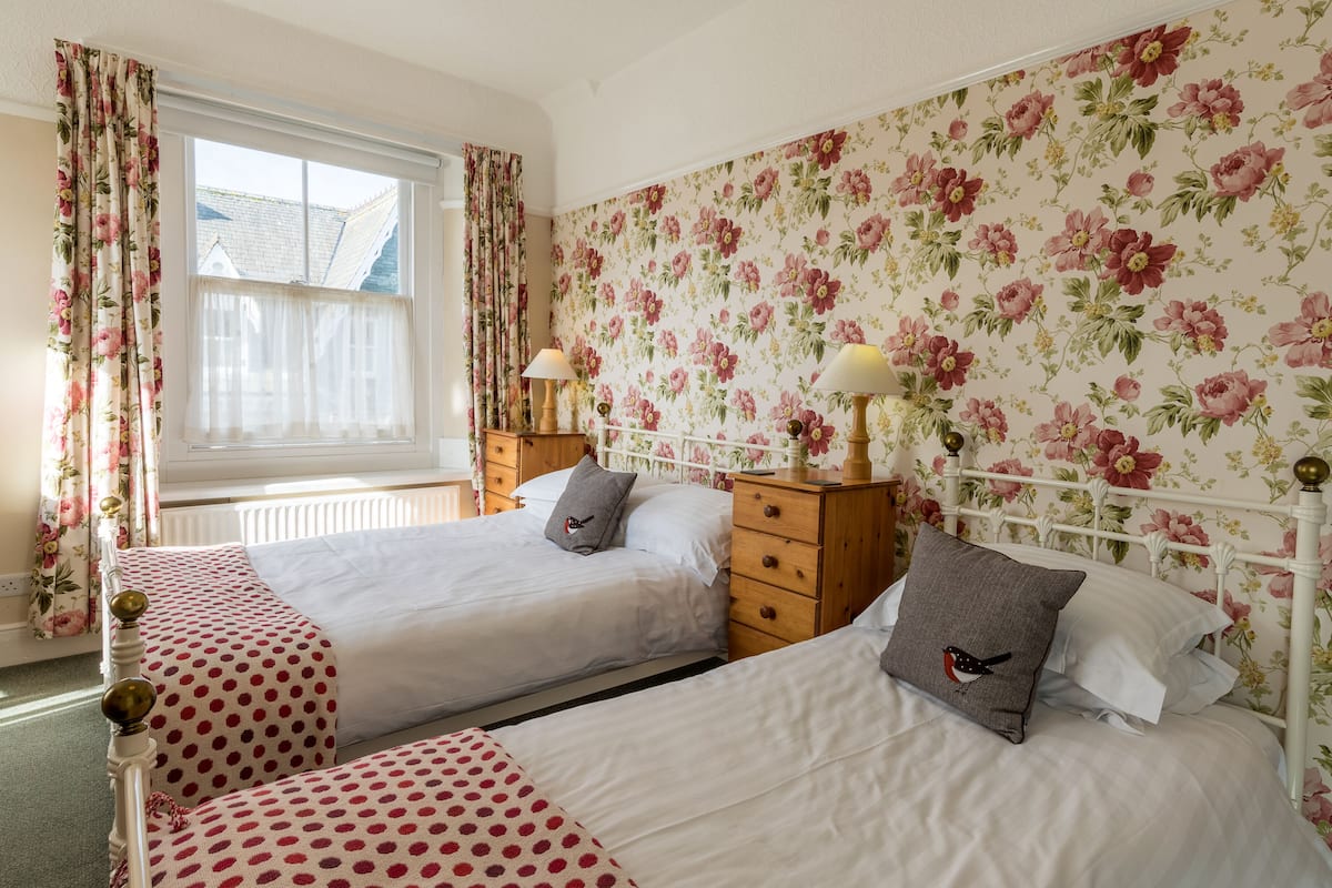 A bedroom with two beds, floral wallpaper, and a window with curtains.
