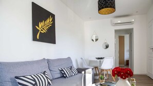 Een moderne woonkamer met een grijze bank, een zwart-wit kunstwerk, een klein tafeltje met boeken en een vaas rode bloemen, en een witte eethoek op de achtergrond.