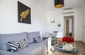 Uma sala de estar moderna com um sofá cinza, uma obra de arte em preto e branco, uma mesinha com livros e um vaso de flores vermelhas, e uma área de jantar branca ao fundo.