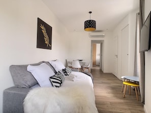 A modern living room with a grey sofa, white fur throw, and a black and white patterned pillow. There is a dining area with a small table and chairs, and a hallway leading to a bedroom.