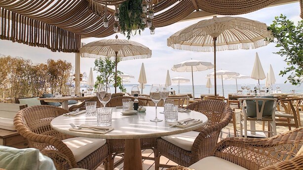 Een buitenplaats met een eethoek, rieten meubilair, ronde tafels en witte parasols.