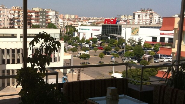 A view from a balcony with a table and chairs, overlooking a parking lot and buildings with various signs, including a Burger King. - Denizhan Otel (Antalya)