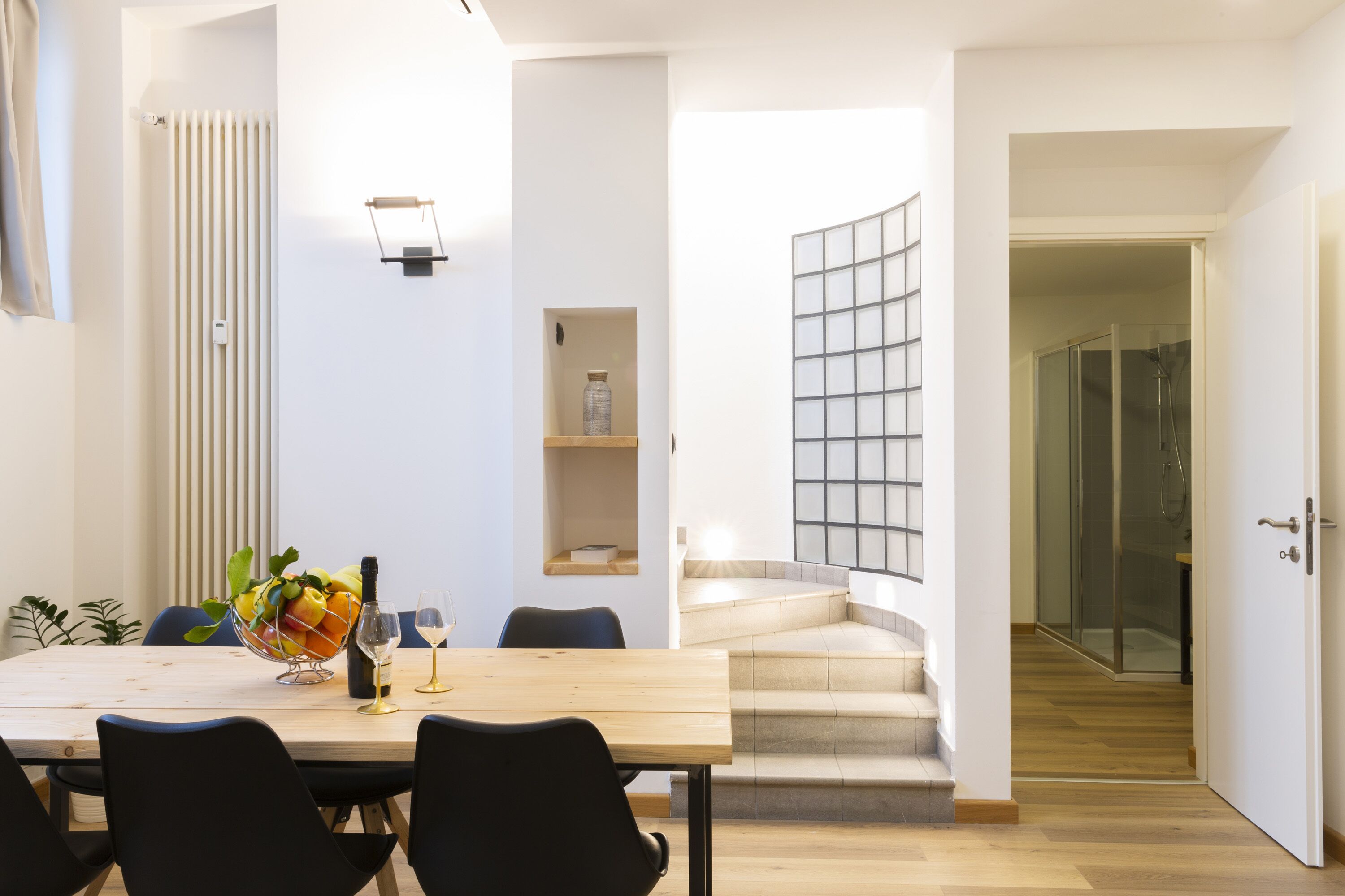 Een moderne eetruimte met een houten tafel, zwarte stoelen en een schaal fruit. Een trap leidt naar een douchecabine met glazen wanden.