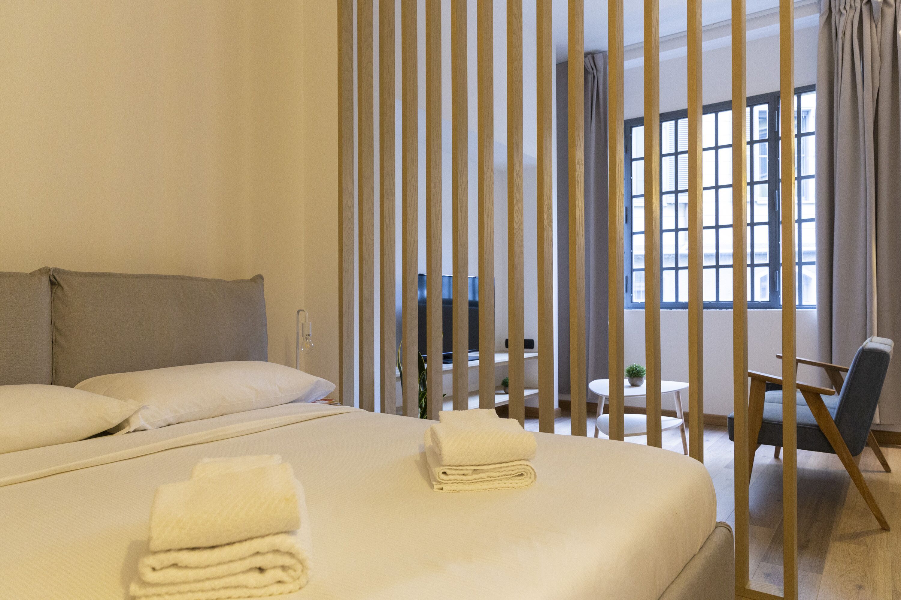 A hotel room with two beds, a desk, a chair, a window with curtains, and a view of buildings outside.