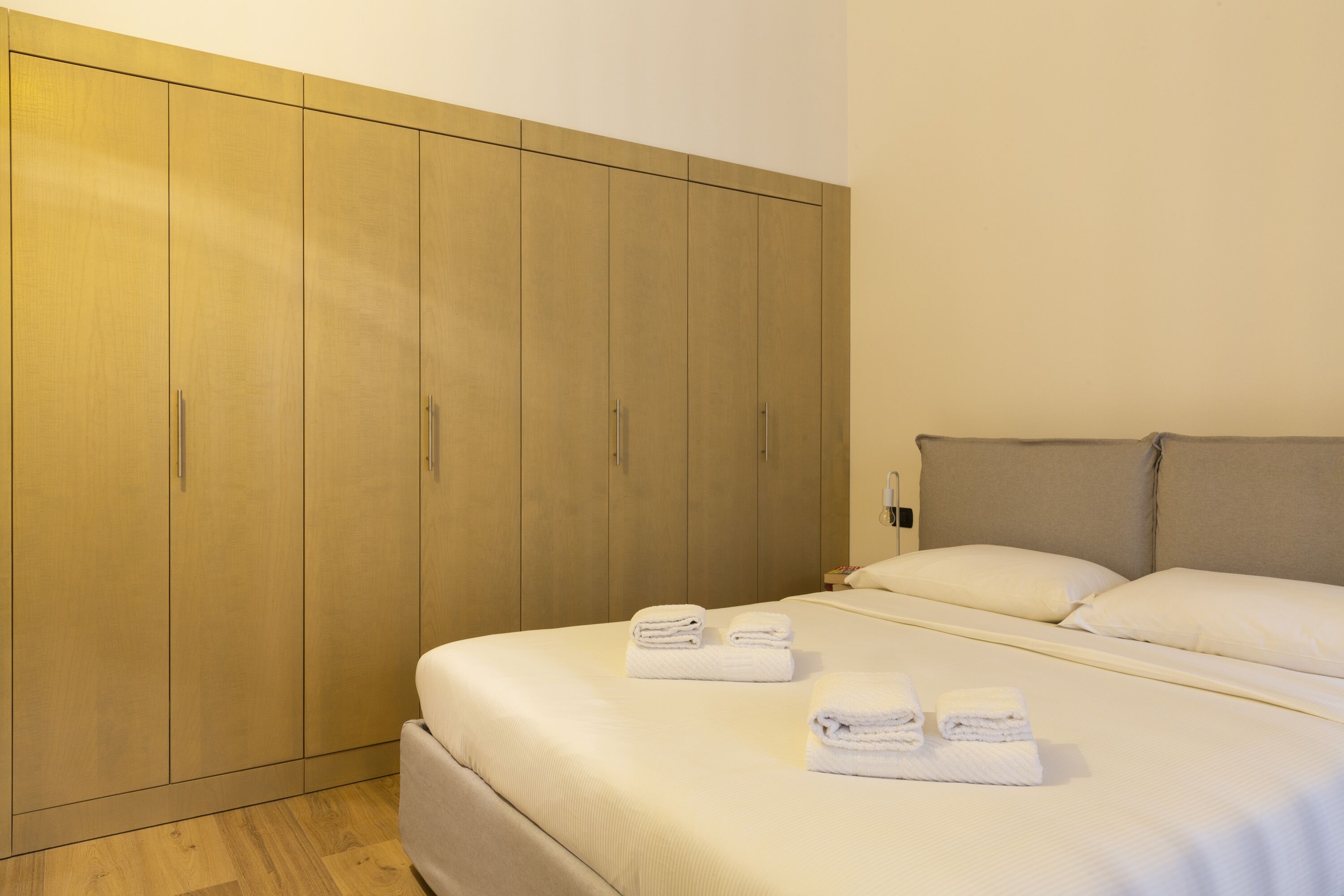 Une chambre d’hôtel dotée d’une grande armoire, d’un lit bien fait avec du linge de lit blanc, et d’une table de chevet avec une lampe.