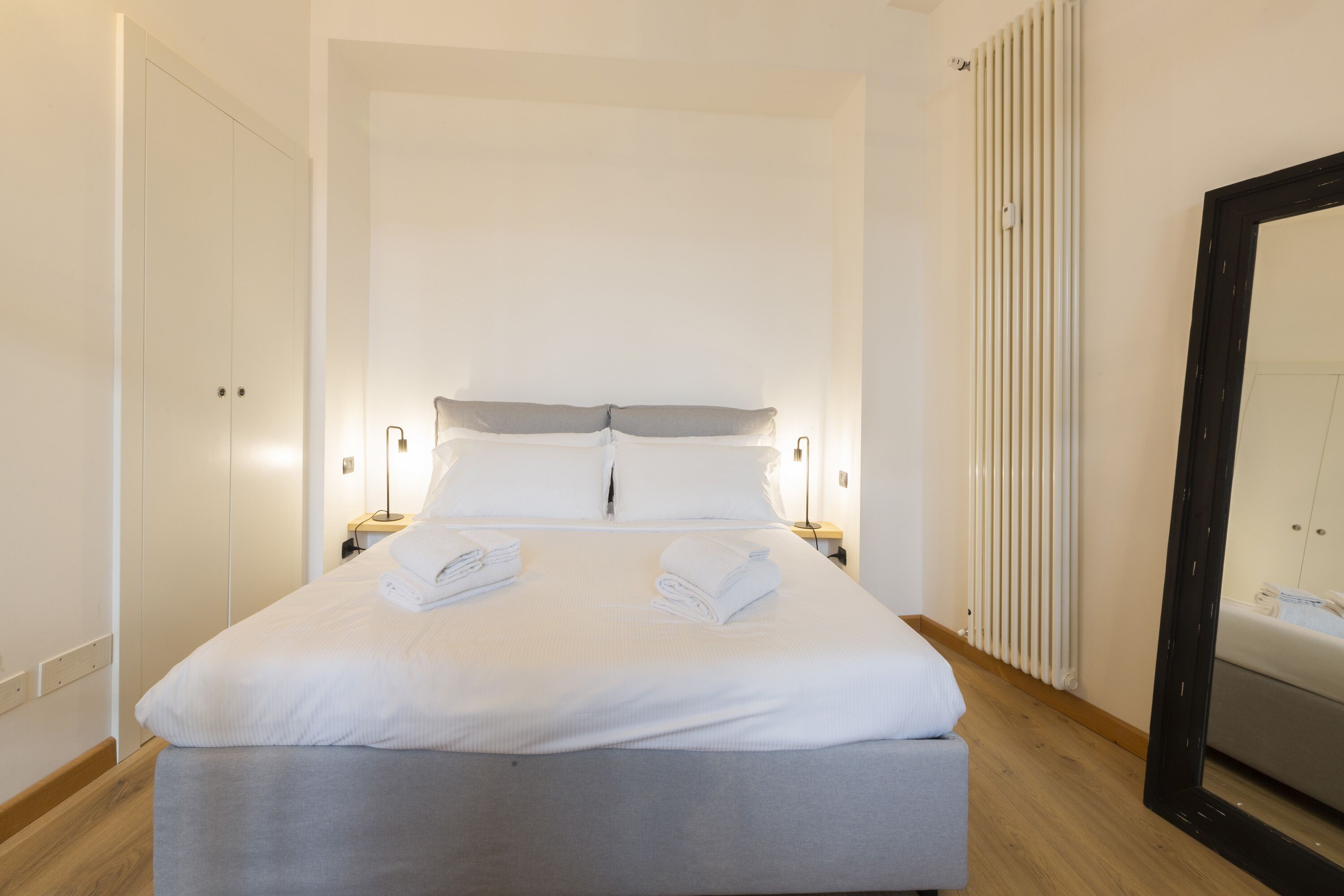 Un lit bien fait avec deux serviettes, une table de chevet avec une lampe, un grand miroir et un radiateur.