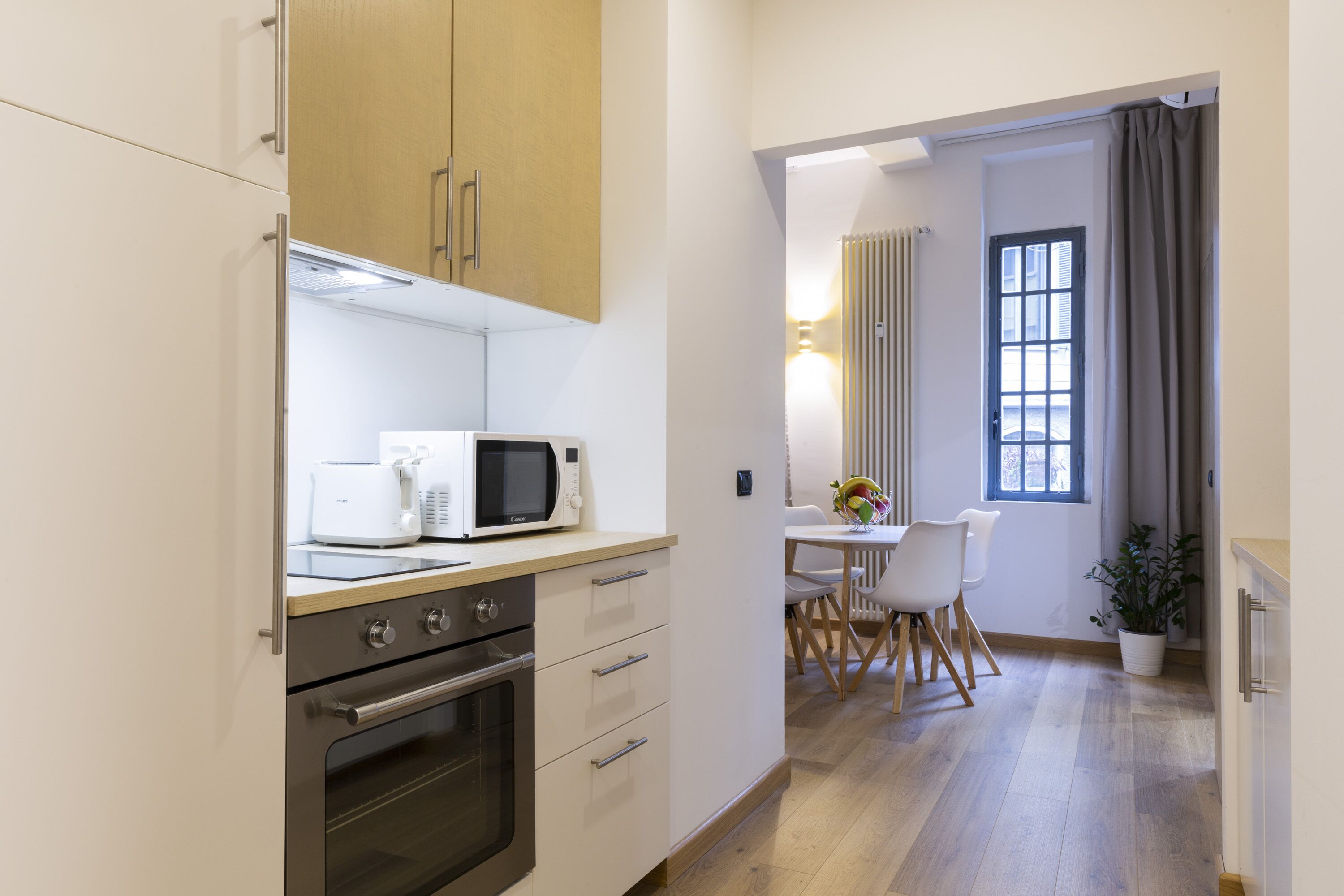 A compact kitchen with a microwave, oven, and wooden cabinets. A dining area with a table and chairs is visible through an open doorway.