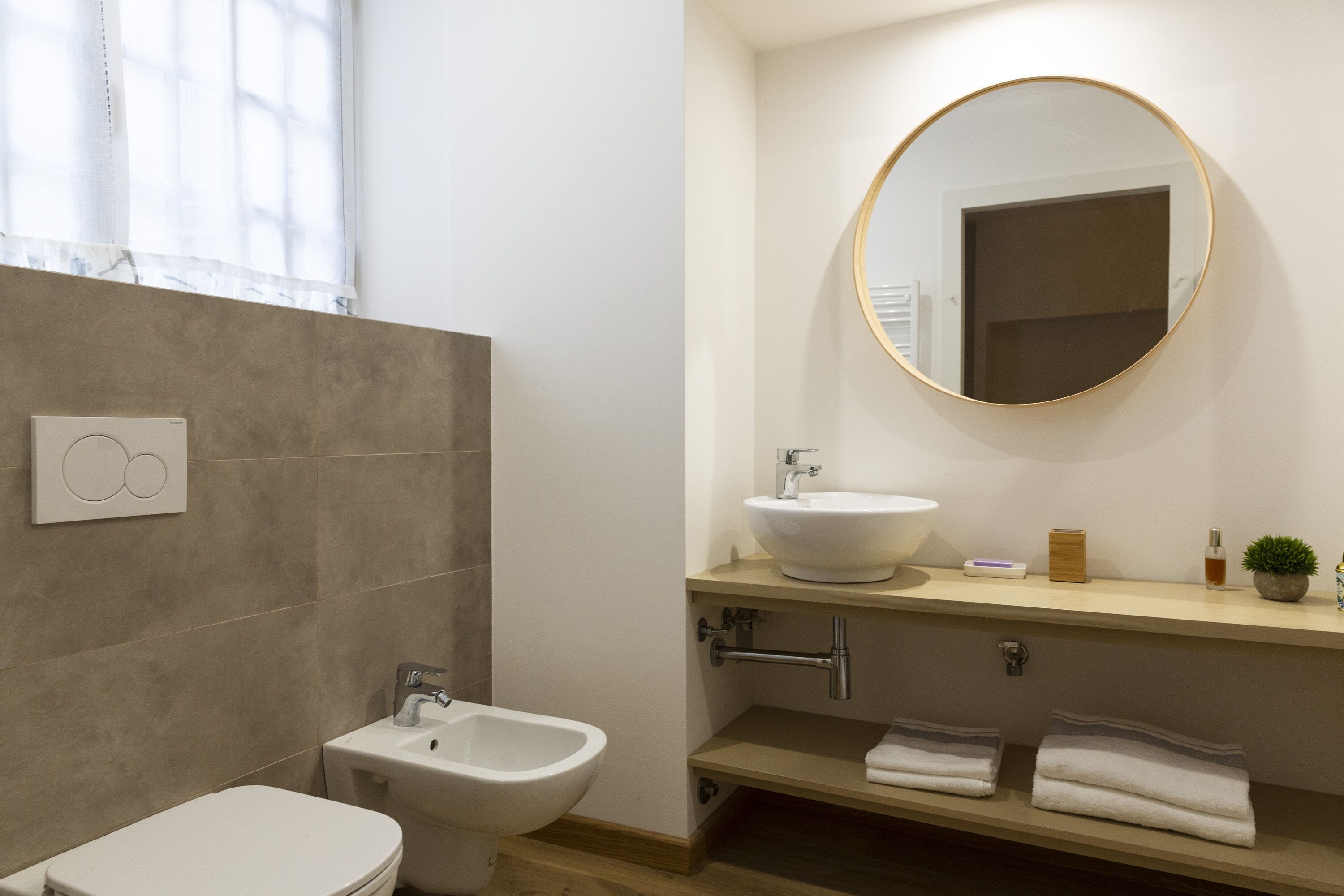 A modern bathroom with a bidet, toilet, sink, and a round mirror.