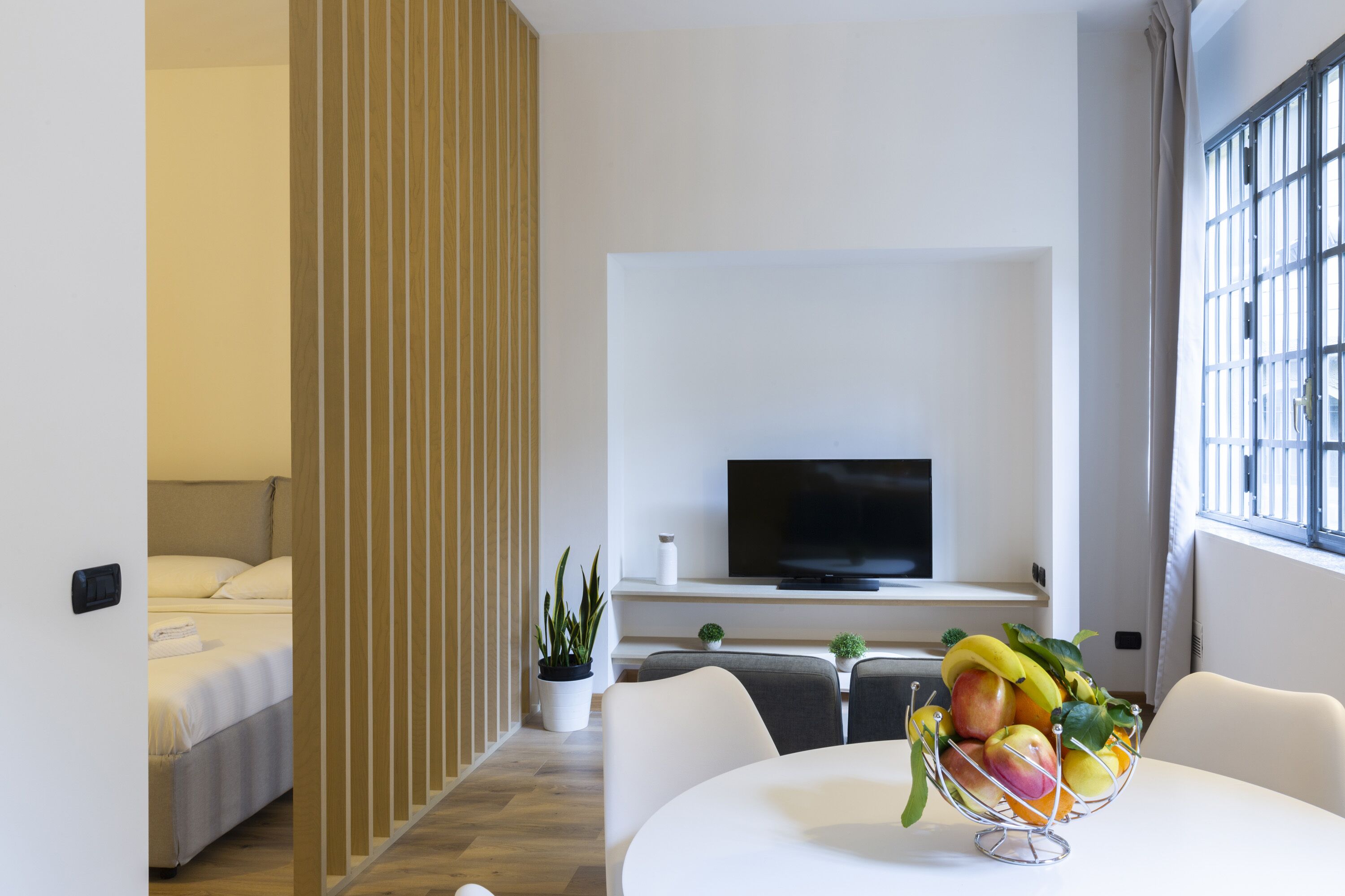 A modern hotel room with a bed, a TV, a dining table with a fruit bowl, and a potted plant.