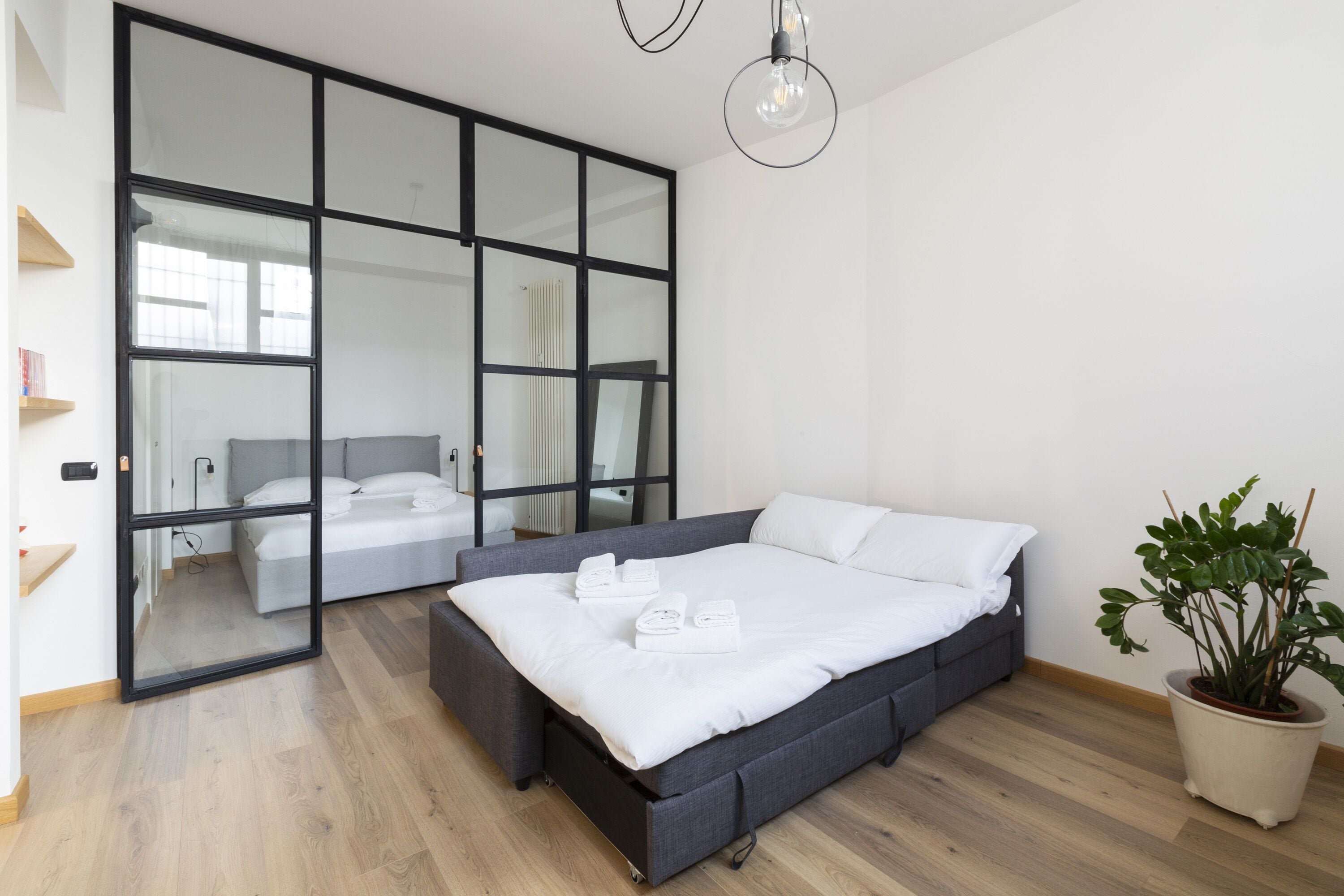 A modern bedroom with a large bed, a potted plant, and a sliding glass door leading to a bathroom.