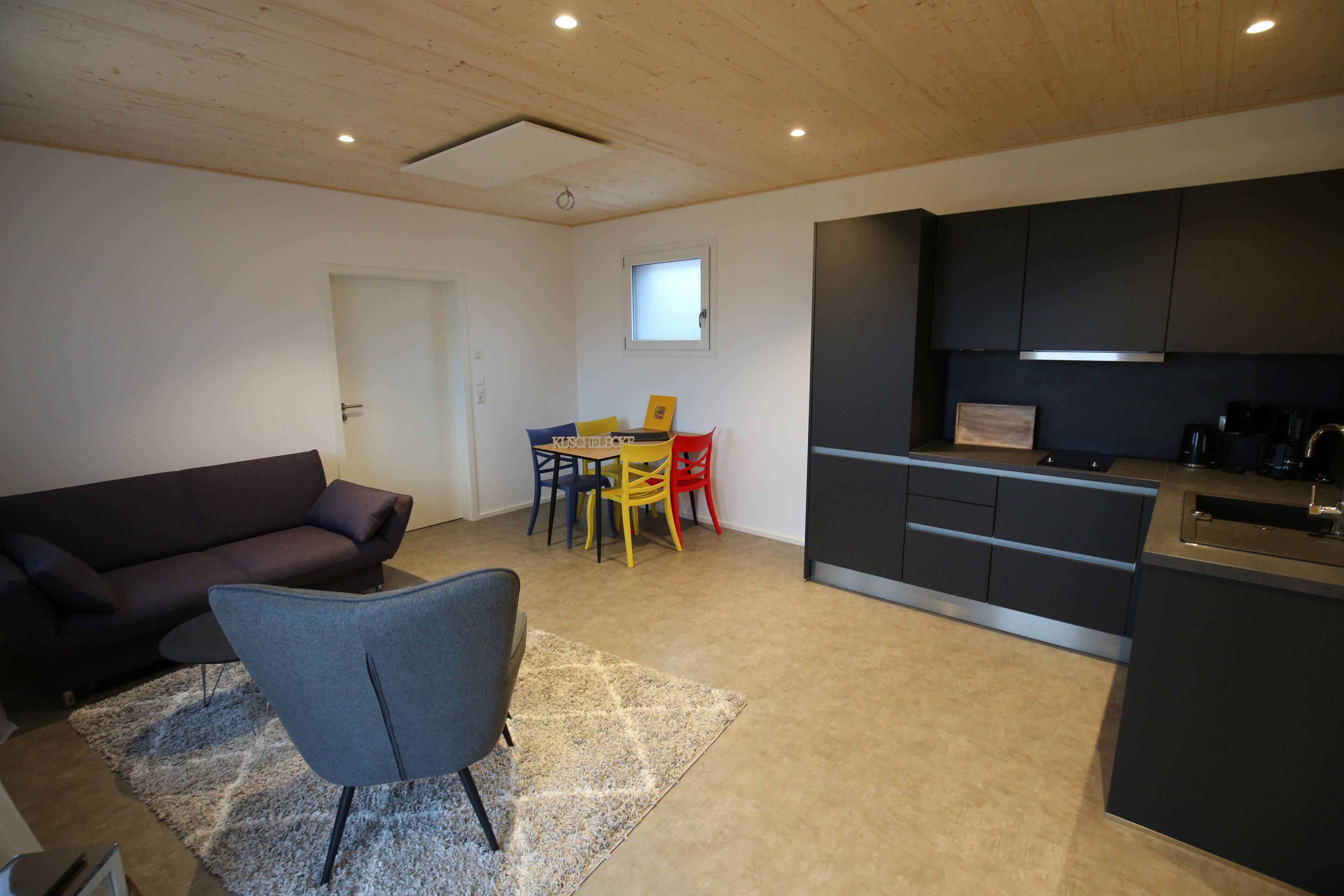 Uma sala de estar moderna com um sofá escuro, uma poltrona cinzenta e uma pequena mesa. É visível, ao fundo, uma área de cozinha com armários escuros e uma janela.