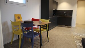 Eine kompakte Küche mit Spüle und Kaffeemaschine, ein Essbereich mit Tisch und bunten Stühlen sowie ein Teppichboden.