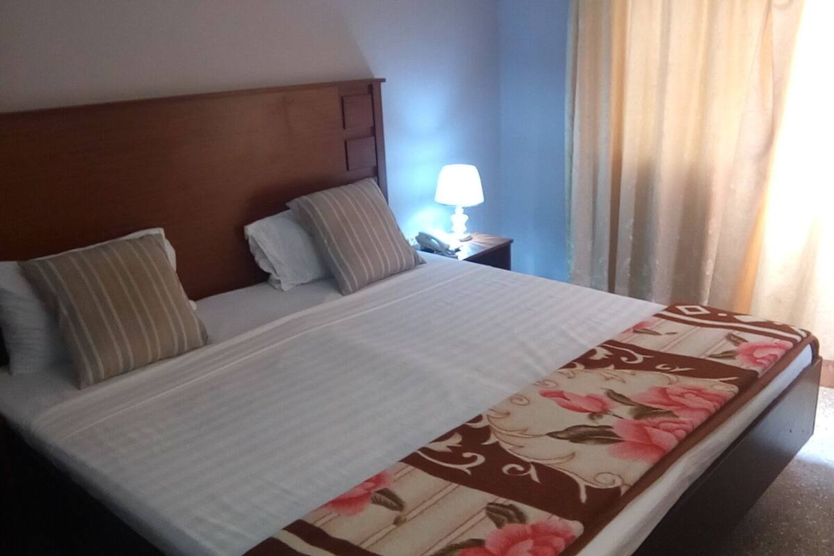 Une chambre d’hôtel avec un lit, une table de chevet, une lampe et une fenêtre avec des rideaux.