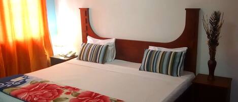 A neatly made bed with floral bedding, a wooden headboard, and striped pillows. There are wall-mounted artworks depicting flowers and a small vase with dried plants on the nightstand.