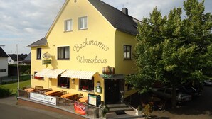 Keltainen rakennus, jonka julkisivussa on teksti ”Beckmann’s Winzerhaus”, ja jossa on ulkotila istumapaikkoineen sekä kyltti, jossa lukee ”Wimmerhaus Urban”.