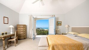 A bedroom with a bed, a desk, a chair, and a dresser. There is a window with a view of the ocean.