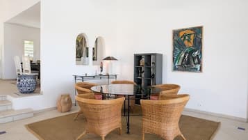 Un comedor con una mesa redonda de vidrio rodeada de sillas de mimbre, una mesita auxiliar de metal negro y un jarrón decorativo.