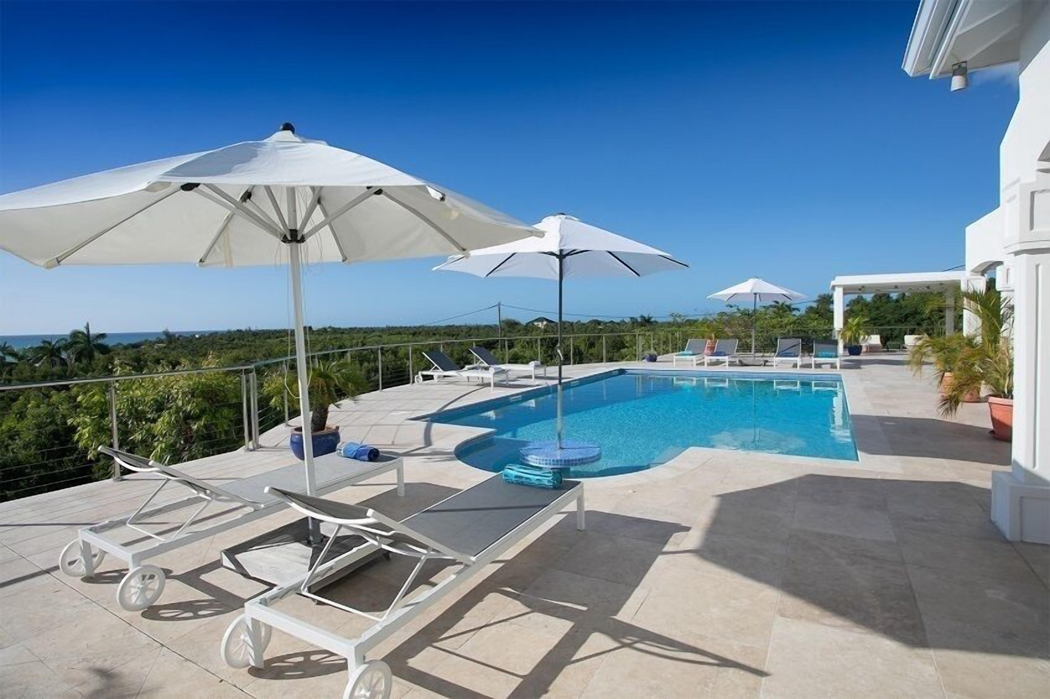 A swimming pool with lounge chairs and umbrellas, surrounded by a white building and greenery.