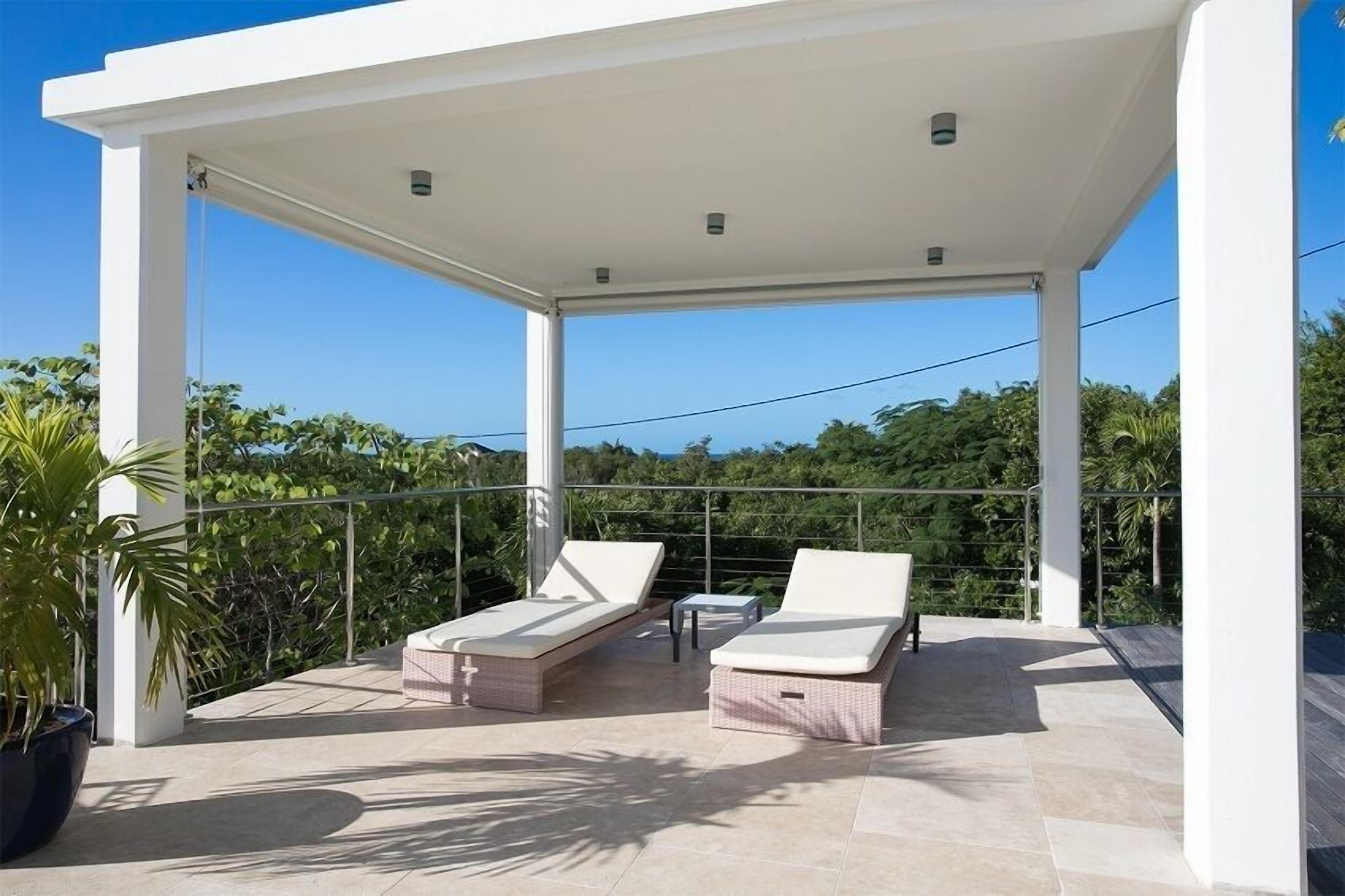 A covered outdoor area with two lounge chairs and a small table, surrounded by a metal railing and potted plants.