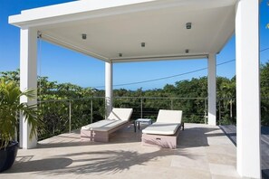 A covered outdoor area with two lounge chairs and a small table, surrounded by a metal railing and potted plants.