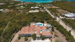 An aerial view of a coastal property with a large house featuring a swimming pool, surrounded by dense vegetation and a clear view of the ocean.
