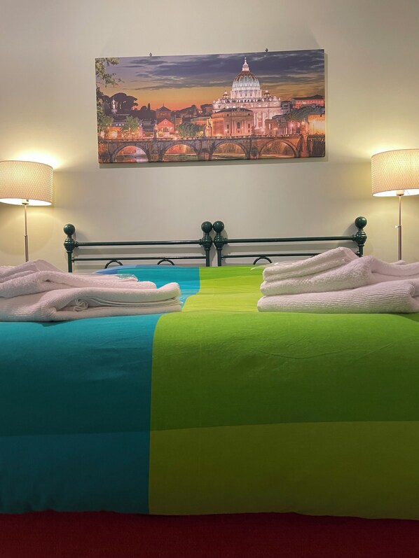 A hotel room with a bed, two bedside lamps, a painting of a cityscape, and a green and blue bedspread.