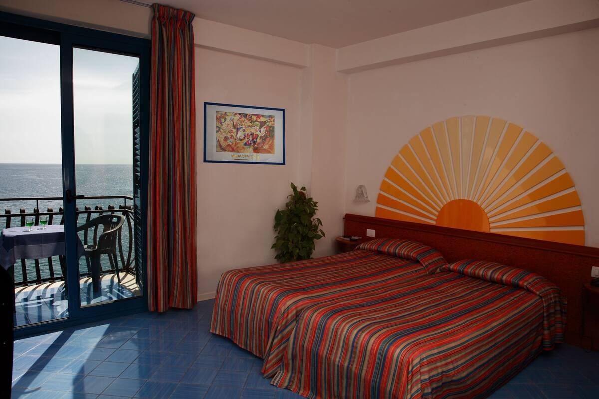 Ein Hotelzimmer mit Bett, Nachttisch, Topfpflanze und Meerblick vom Balkon.