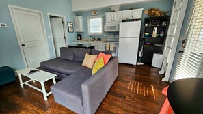 A compact living space with a grey sofa, a small white table, a kitchen with white cabinets, a microwave, and a refrigerator.
