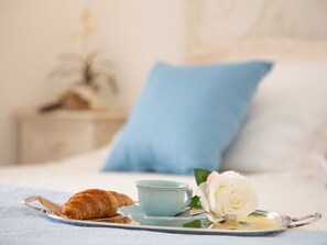Un plateau avec un croissant, une tasse bleue et une rose blanche, posés sur une table de chevet avec un oreiller bleu et une plante.