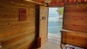 A wooden cabin room with a bed, a small table, and a door leading to a balcony with a view of the ocean. -  Paradise Over the Water Cabins in San Blas (Narganá)