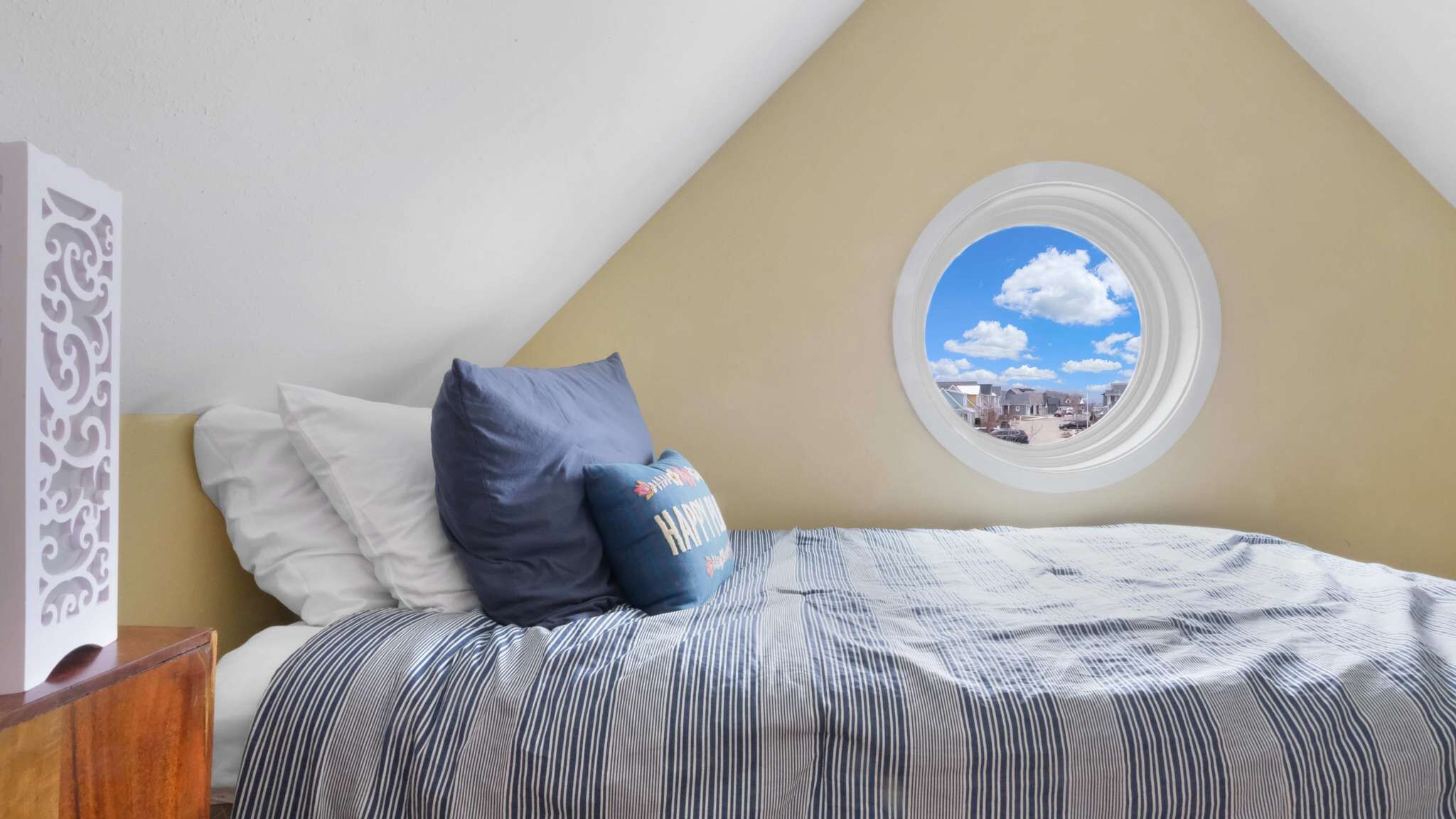 A bedroom with a sloped ceiling, a window with a view of a residential area, a bed with striped bedding, and decorative pillows.