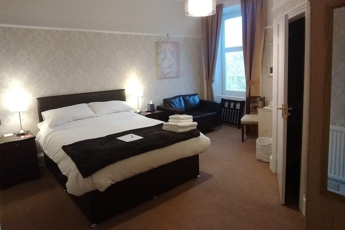 A hotel room with a large bed, bedside lamps, a black leather sofa, a radiator, and a door to the right.